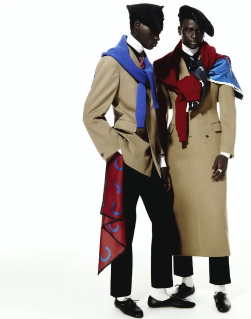 Two dark-skinned men model beige overcoats and black berets in a studio, accessorized with colorful scarves, set against a plain white backdrop.