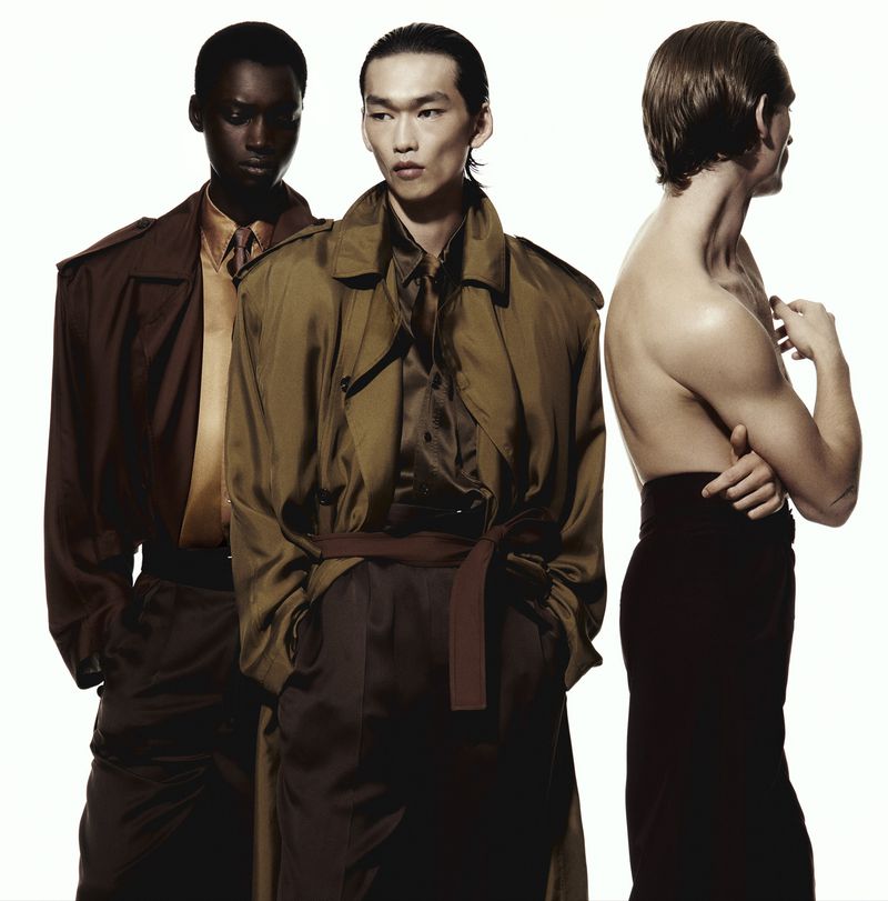 Three diverse men showcased in a studio setting. One shirtless man stands in the back, while two other men wear satin shirts and trench coats.