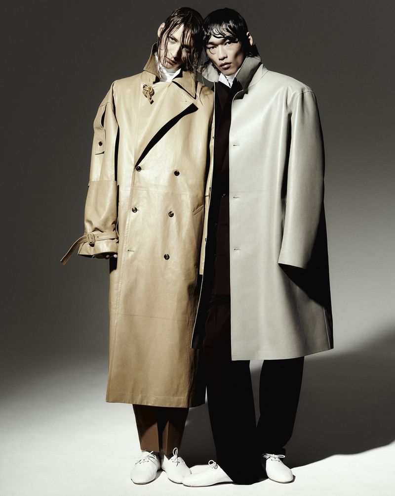 Two fashion models in neutral tone trench coats pose in a studio. One model is of Asian descent; the other appears to be of European descent.