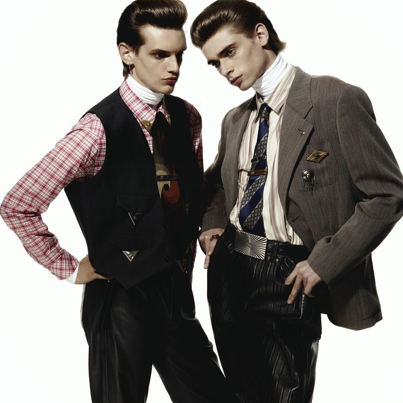 Two adult male fashion models in patterned shirts, vests, and suits pose on a white background in a studio environment for a portrait.