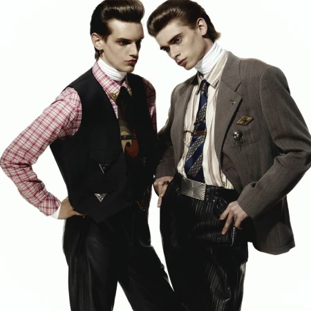 Two adult male fashion models in patterned shirts, vests, and suits pose on a white background in a studio environment for a portrait.