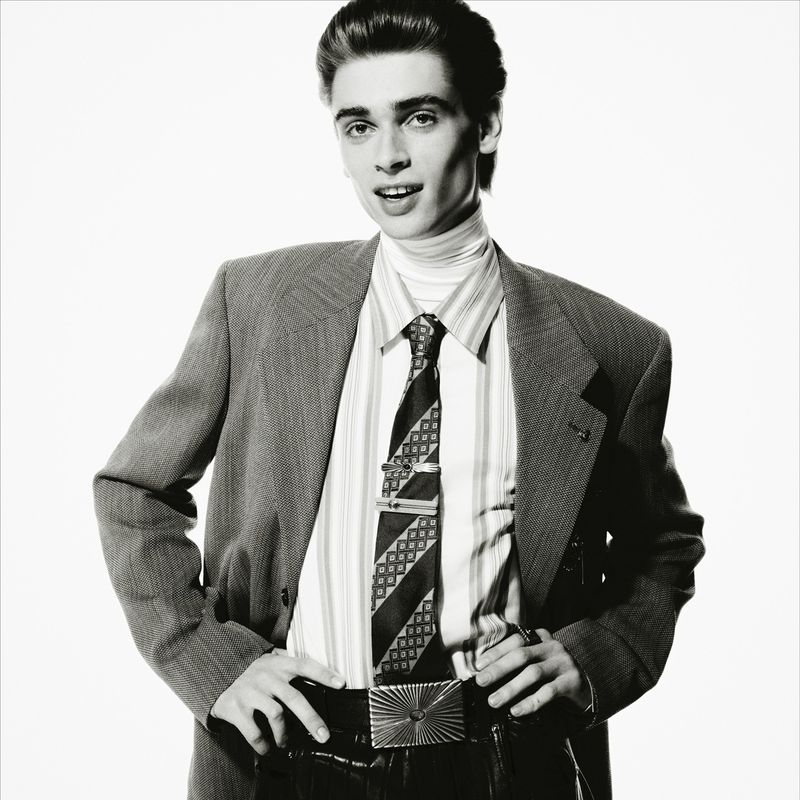 Portrait of a young adult male in a suit with a patterned tie and shiny belt buckle against a bright white background.