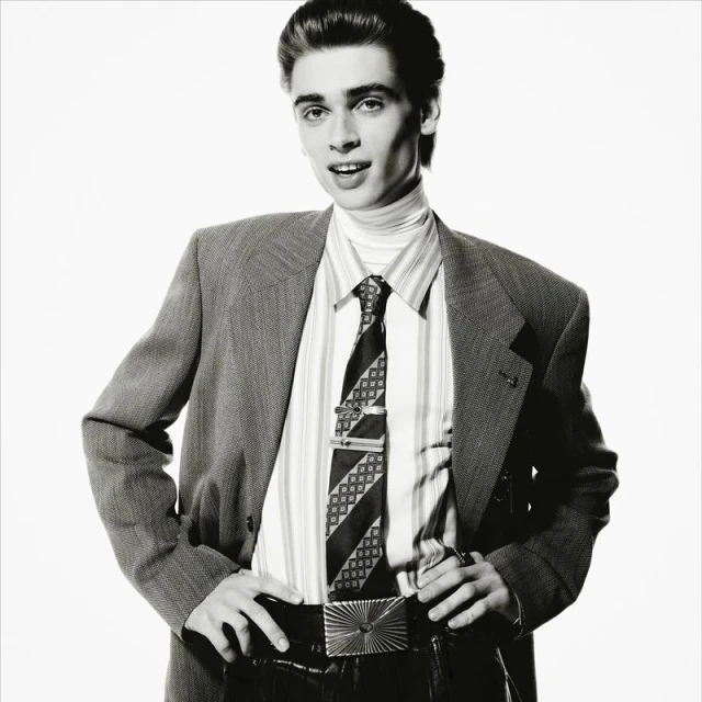 Portrait of a young adult male in a suit with a patterned tie and shiny belt buckle against a bright white background.
