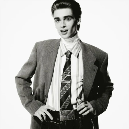 Portrait of a young adult male in a suit with a patterned tie and shiny belt buckle against a bright white background.