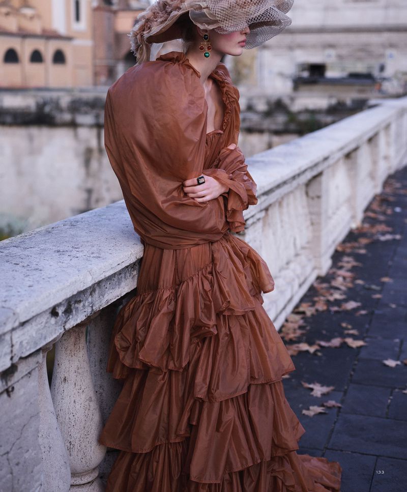 Fashion shot of woman in brown tulle gown and veil hat, leaning on stone ledge with city and building background.