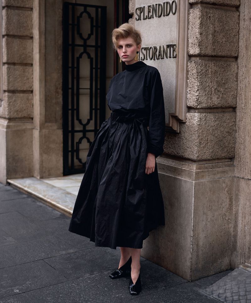 A blonde fashion model in a black dress poses against a building with a restaurant sign in an outdoor urban setting.