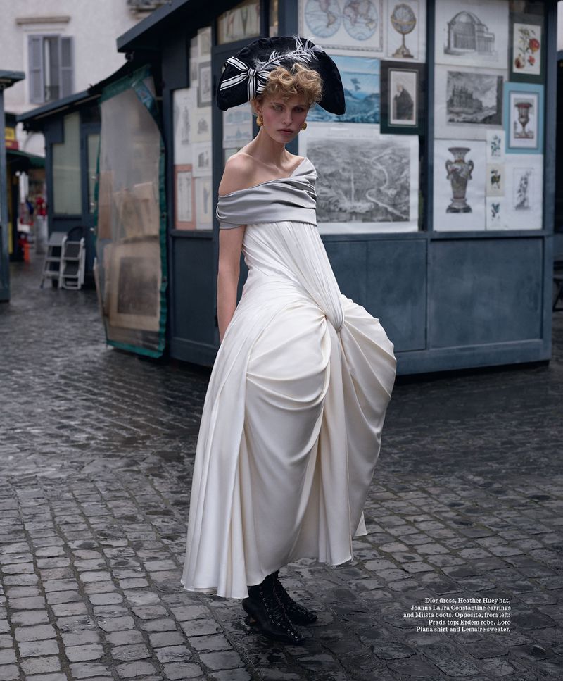 A fashion-forward woman in a white gown and striking hat stands in a European street, showcasing high-fashion editorial style.