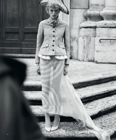 Fashion editorial shot in black and white featuring a blonde model in a skirt, blazer, heels, and hat posing on stairs.