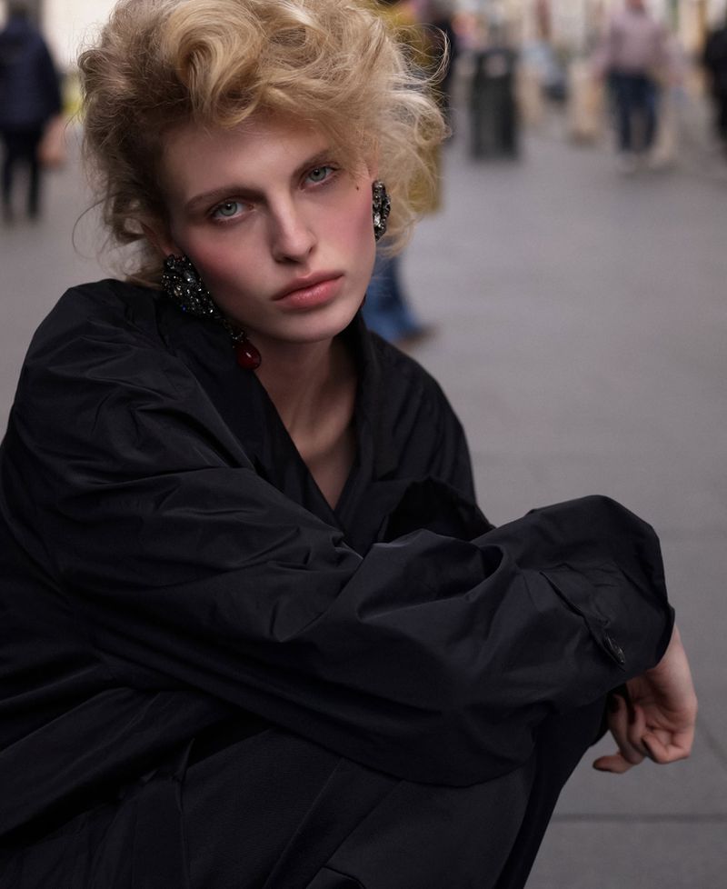 Woman with blond curly hair, dark jacket, and large earrings in an outdoor setting with blurred figures, shallow depth of field.