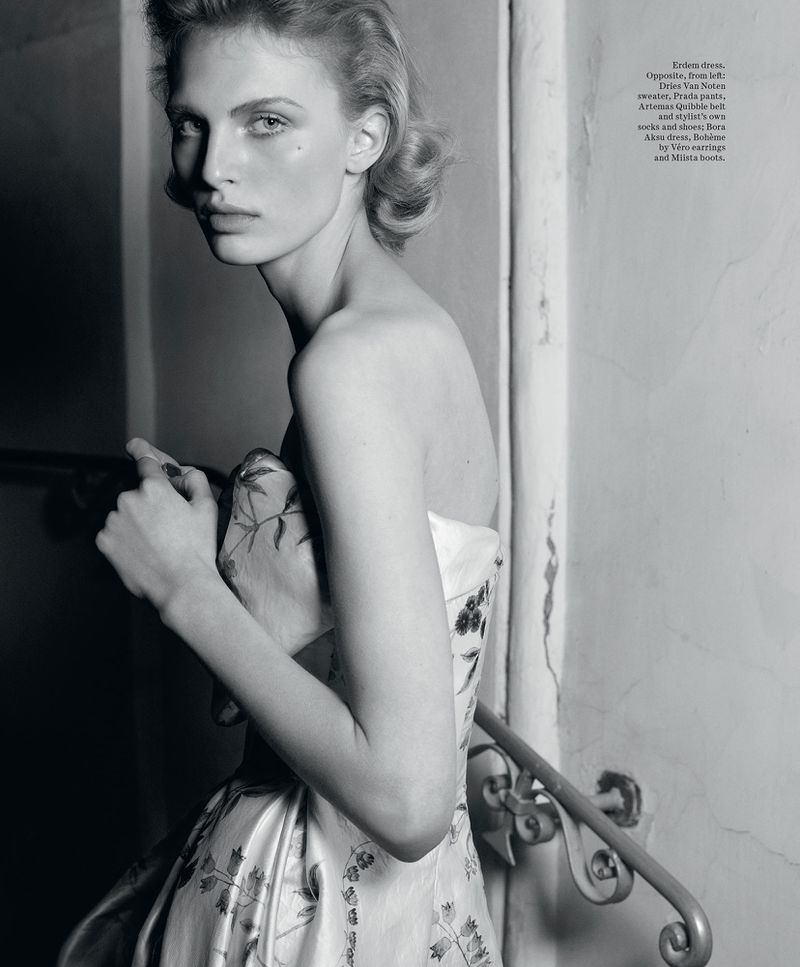 Black and white fashion shot of a young female model in a floral gown near a staircase, posing with a neutral expression.