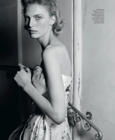 Black and white fashion shot of a young female model in a floral gown near a staircase, posing with a neutral expression.