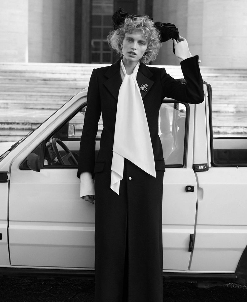 Black and white fashion shot of a female model in a black suit with a white tie, standing in front of a white car and stone steps.