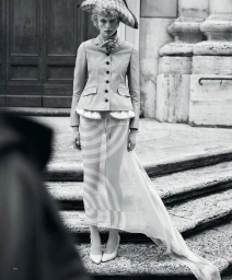 Model standing on stone steps wearing a tailored jacket, patterned sheer skirt, hat, and heels against a building exterior.