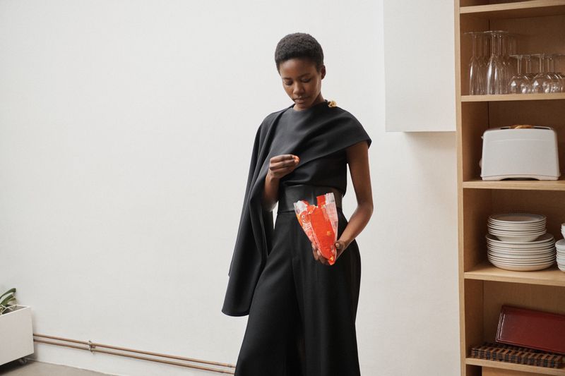 Elegant Black Woman in Chic Outfit Holding a Red Object in an Indoor Setting with White Walls and a Wooden Shelf Displaying Dishes.