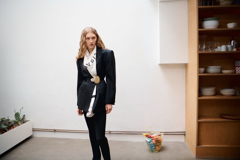 A fair-skinned adult woman with wavy hair poses indoors wearing a black blazer with an oversized buckle and black pants, fashion editorial.