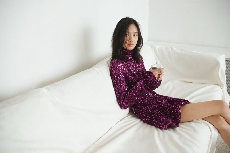 A stylish young woman relaxes on a white couch in a sparkly purple dress, holding a clear glass. Minimalist modern interior aesthetic.
