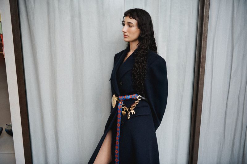 Adult female in black blazer dress with colorful belt poses in an indoor environment with natural light; fashion forward and elegant.