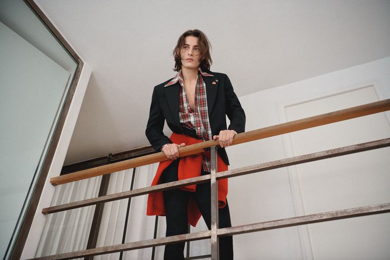 Low-angle view of adult male fashion model posing indoors with plaid shirt, jacket, and red sweater tied around his shoulders.