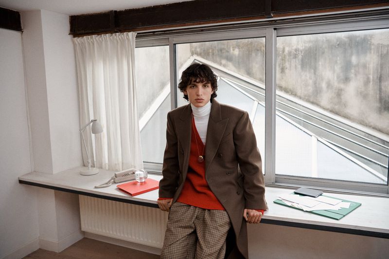A young adult stands indoors by a large window wearing a brown coat over a red sweater and plaid pants, next to a desk with office supplies.
