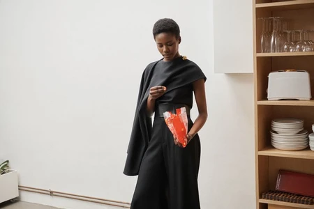 Model in black top and trousers holding a red package inside a room with a wooden shelving unit