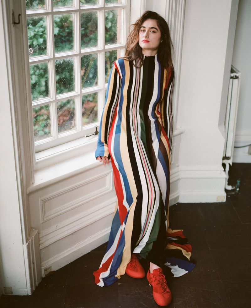 Indoor shot of a young woman in a multicolored striped dress leaning against a window with red sneakers, posing for fashion content.