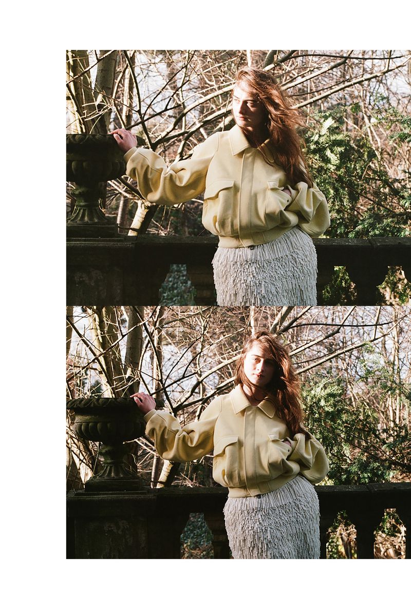 Young woman in a yellow bomber jacket and fringe skirt poses on a stone balcony overlooking a park with trees on a sunny day.