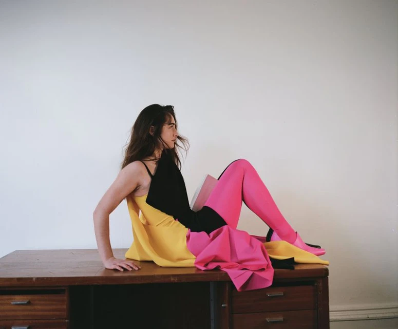 A young woman with long brown hair is seated on a brown wooden desk, posing with colourful yellow, pink, and black fabrics against a white wall.