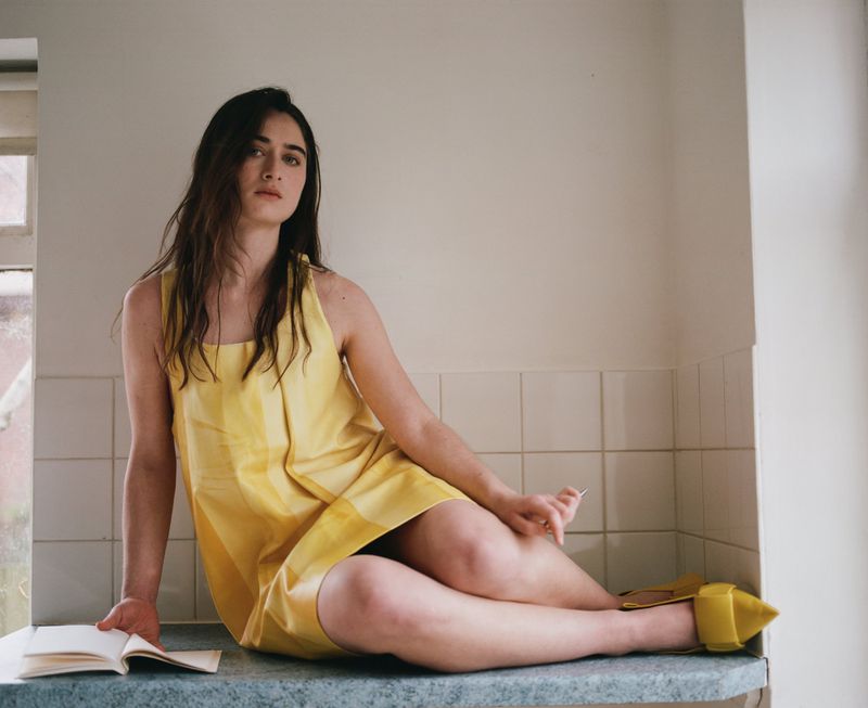 Young woman in yellow dress sits on countertop in domestic kitchen reading an open book in natural light. Soft, desaturated colors.