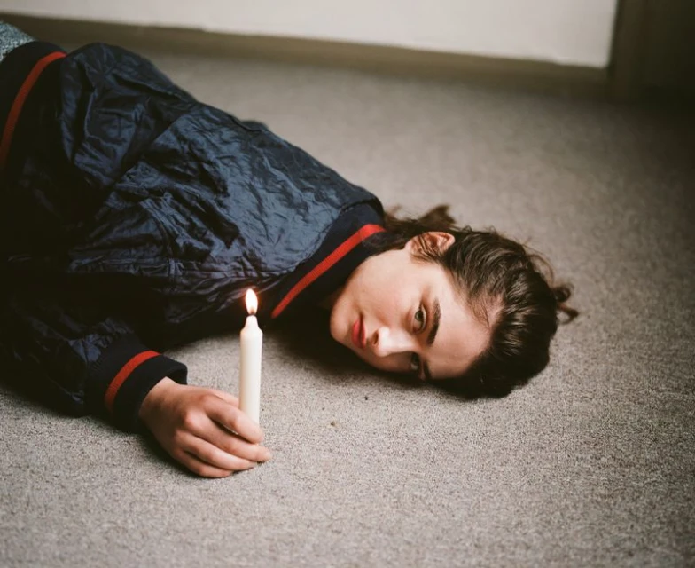 A young woman lies on the floor holding a lit white candle, wearing a blue jacket with red trim, against a neutral wall in soft lighting.