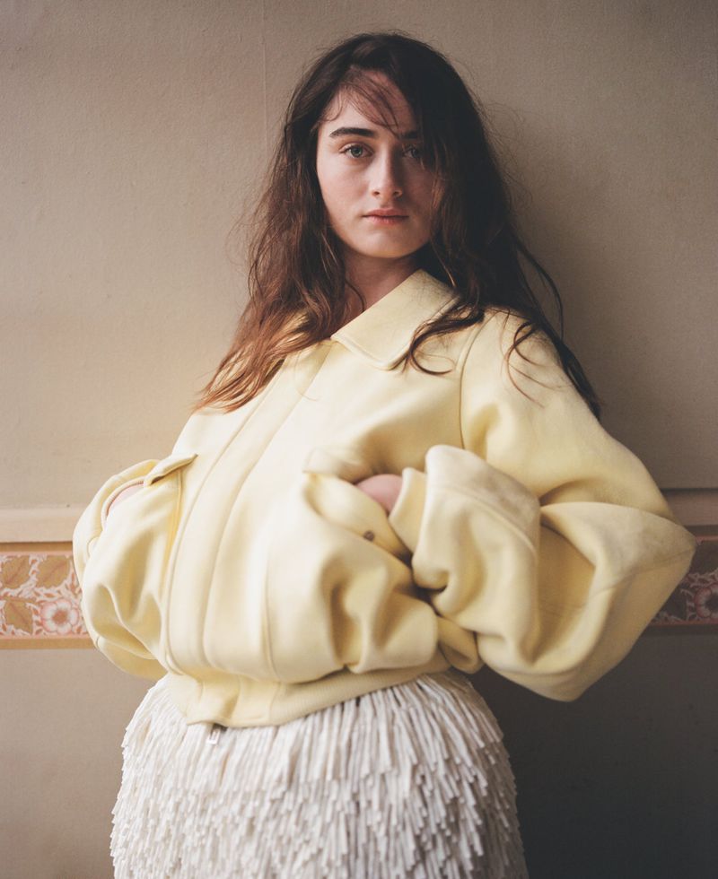 Portrait of a young adult woman with long brown hair wearing a light yellow jacket and white fringed skirt, posing indoors against a wall.