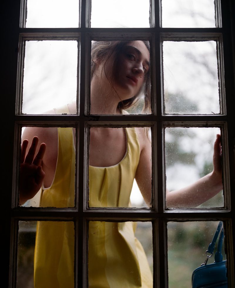 Woman in yellow dress stands behind a paneled window, hand pressed against glass, looking wistfully to the side, with a blue bag visible.