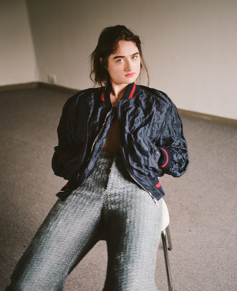 Portrait of a young woman in a navy bomber jacket and sparkly silver trousers sitting on a stool in a room with minimal decoration.