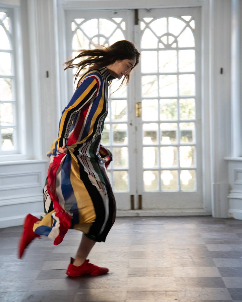 Fashionable woman runs in a striped dress and red shoes in an ornate room with large white doors and checkered floor tiles.