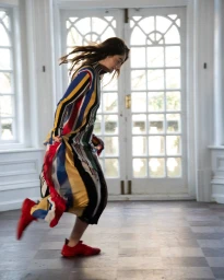Actor Raffey Cassidy in a colourful striped dress and red sneakers dancing indoors before large double doors
