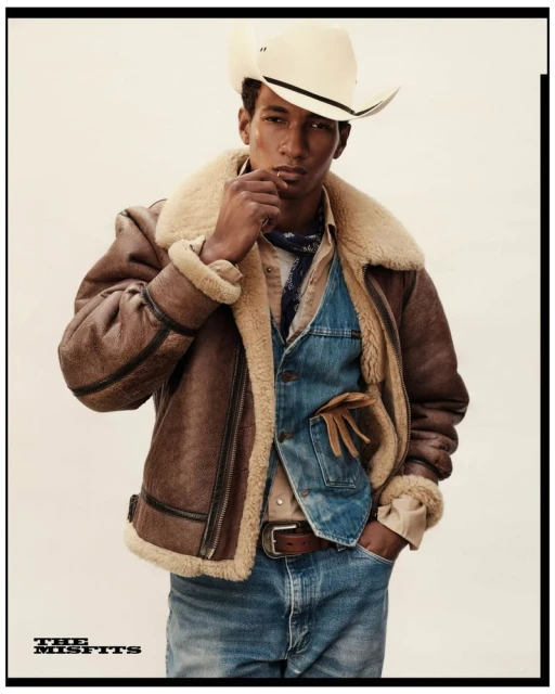 Man in cowboy hat and shearling jacket poses in front of off-white backdrop. Jeans and denim vest create a timeless fashion statement.