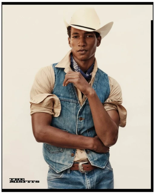 Portrait of a Black male model in a western style with a cowboy hat, denim vest, and jeans against a neutral background in a studio.