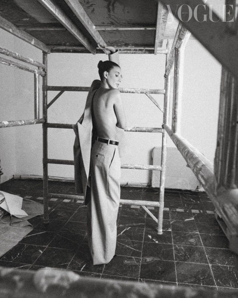 Black and white portrait of a model in trousers, standing within scaffolding. Minimalist composition with a vintage Vogue logo.
