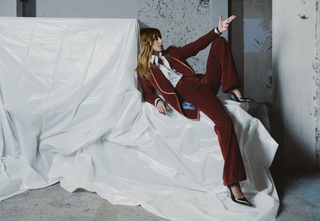 A stylish woman in a burgundy suit reclines on a draped white surface, exuding confidence in a raw, urban setting with textured walls.
