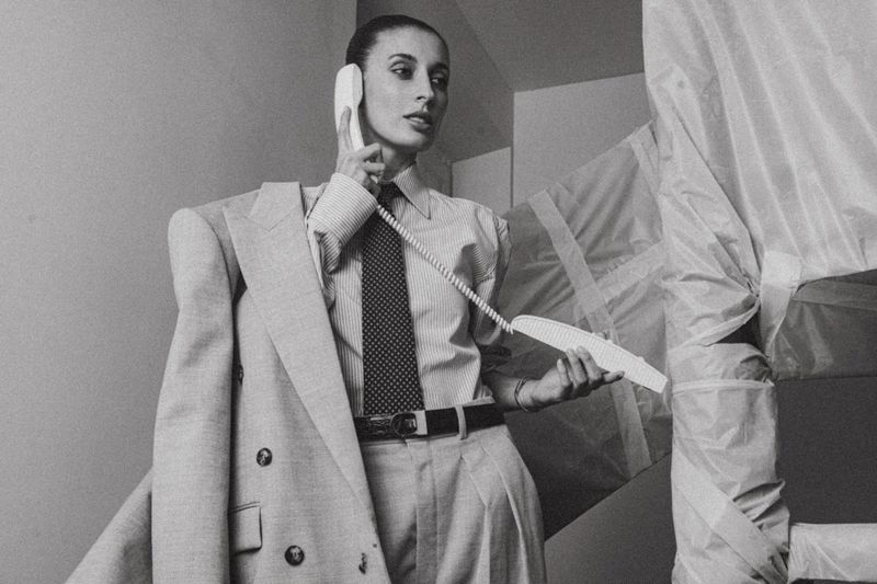 Monochrome portrait of woman in pinstripe shirt, tie, and suit jacket holding telephone receiver indoors, surrounded by covered objects.