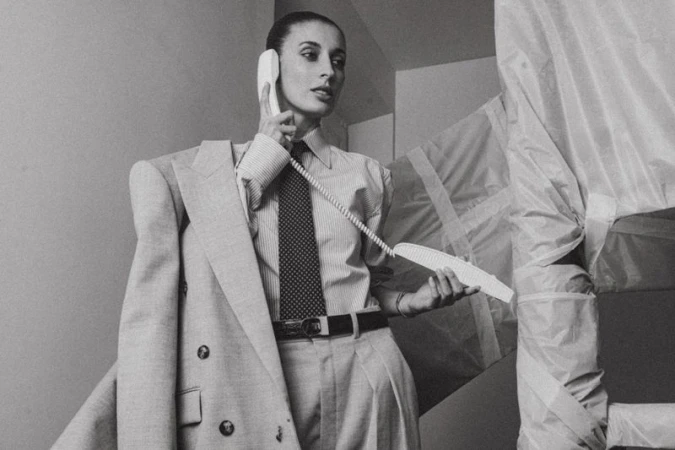 Monochrome portrait of woman in pinstripe shirt, tie, and suit jacket holding telephone receiver indoors, surrounded by covered objects.