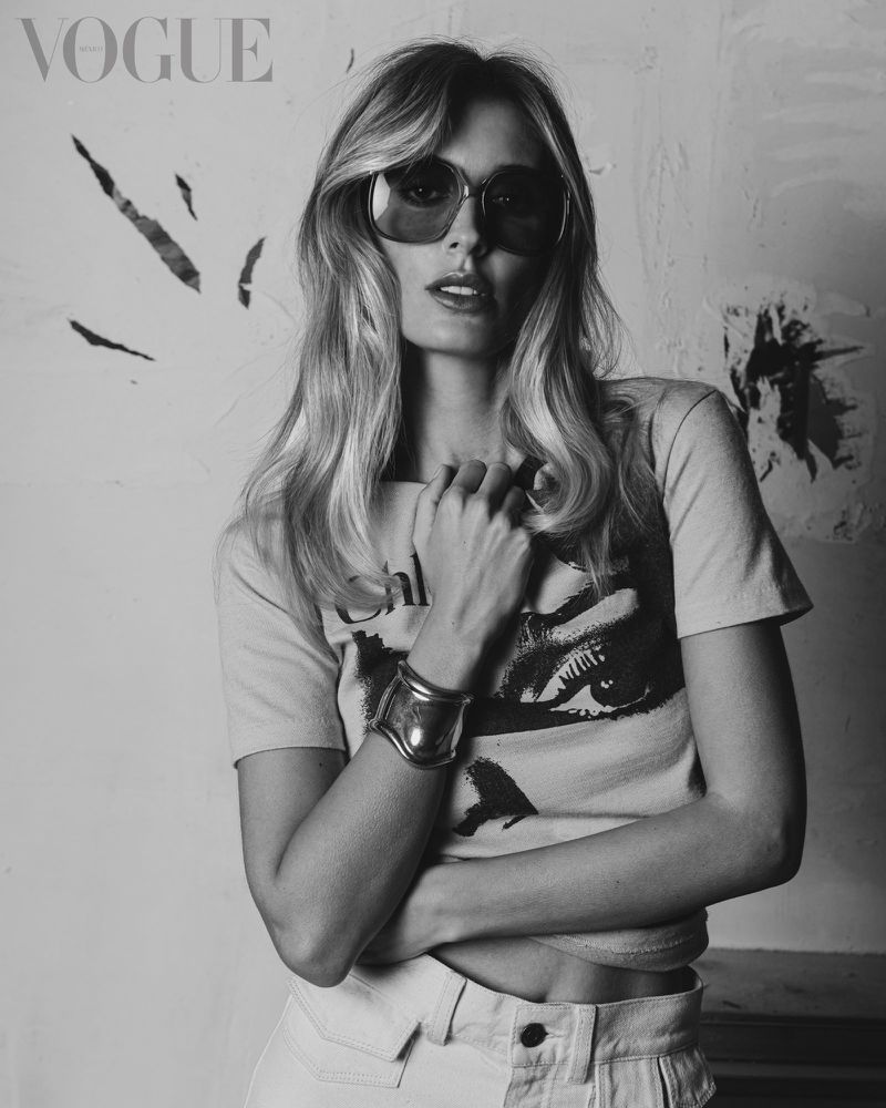 Black and white portrait of a blonde woman wearing sunglasses, a t-shirt, and jeans while posing in front of a textured wall.