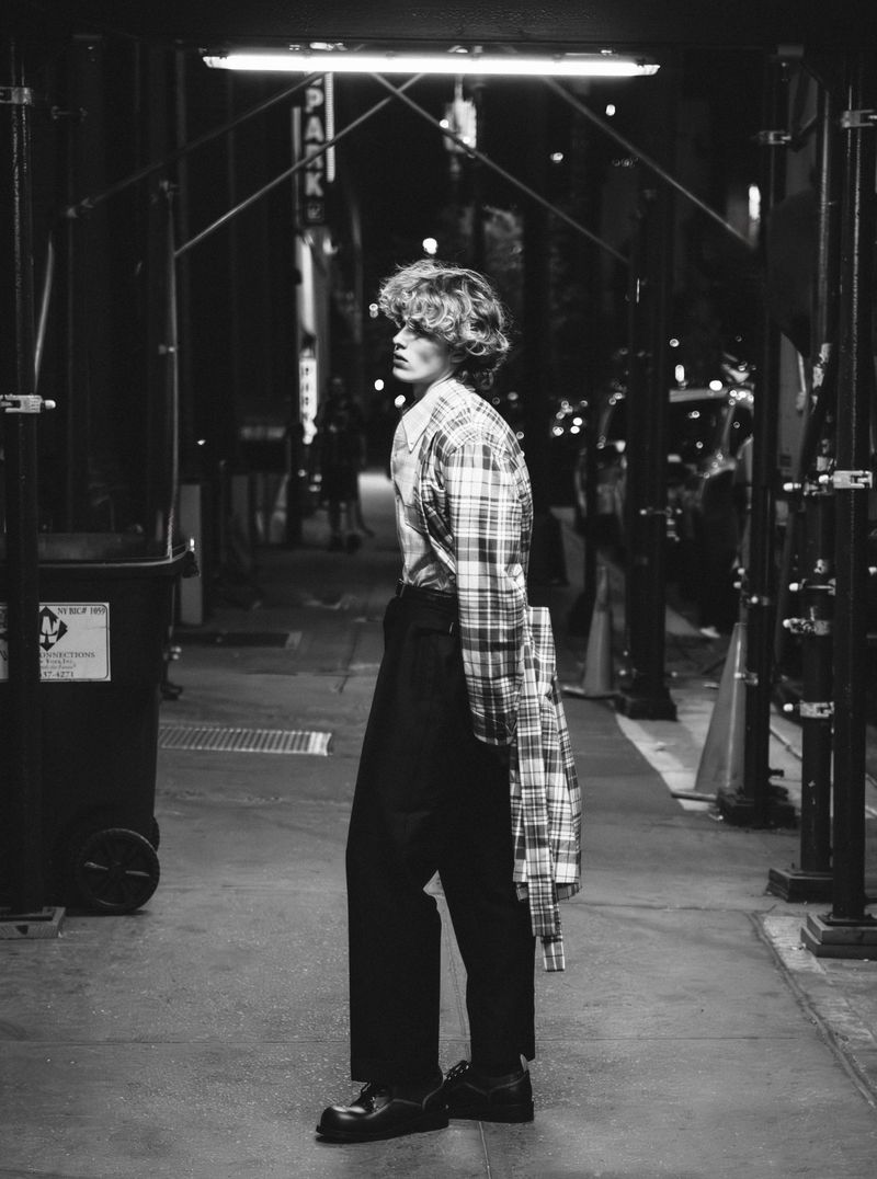 Monochrome portrait of a young adult male fashion model with curly hair in a plaid shirt walking on a city street at night.