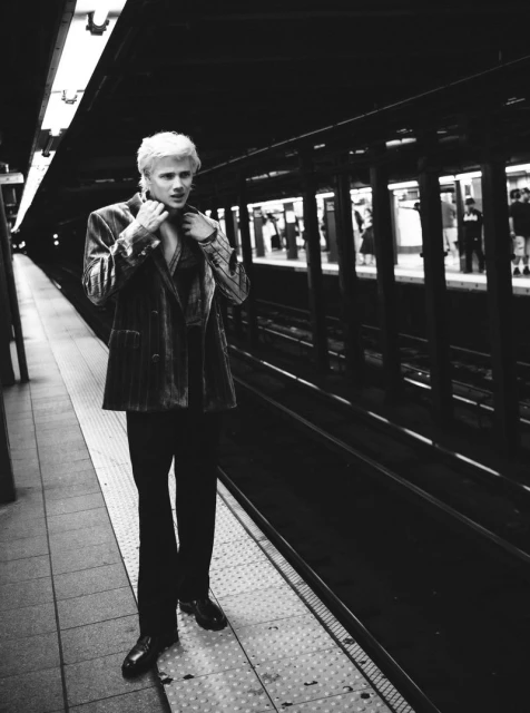 Black and white portrait of an androgynous person in a suit jacket at a subway station, waiting at the platform edge.