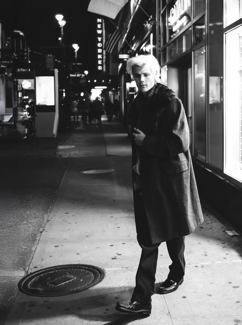 Black-and-white street style photo of a blonde male model in a coat and dress pants walking on a sidewalk at night with city lights.