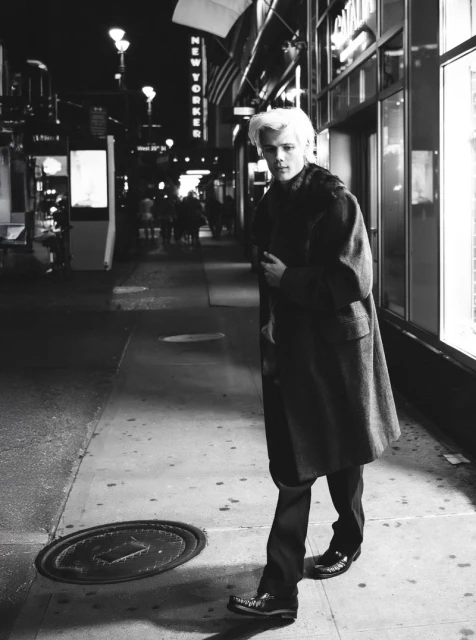 Black-and-white street style photo of a blonde male model in a coat and dress pants walking on a sidewalk at night with city lights.