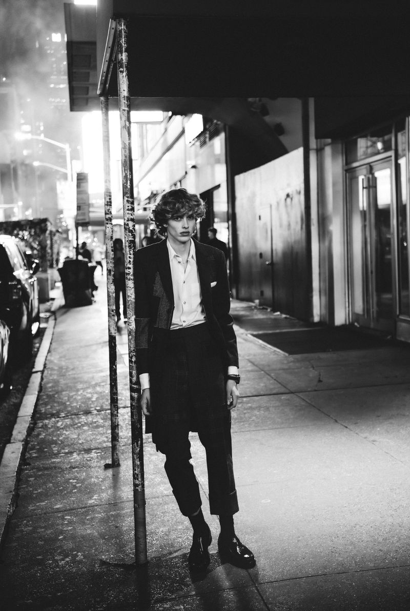 Black and white portrait of a male model in a suit leaning on a pole on a city sidewalk at night with buildings in the background.