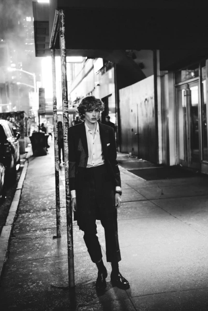 Black and white portrait of a male model in a suit leaning on a pole on a city sidewalk at night with buildings in the background.