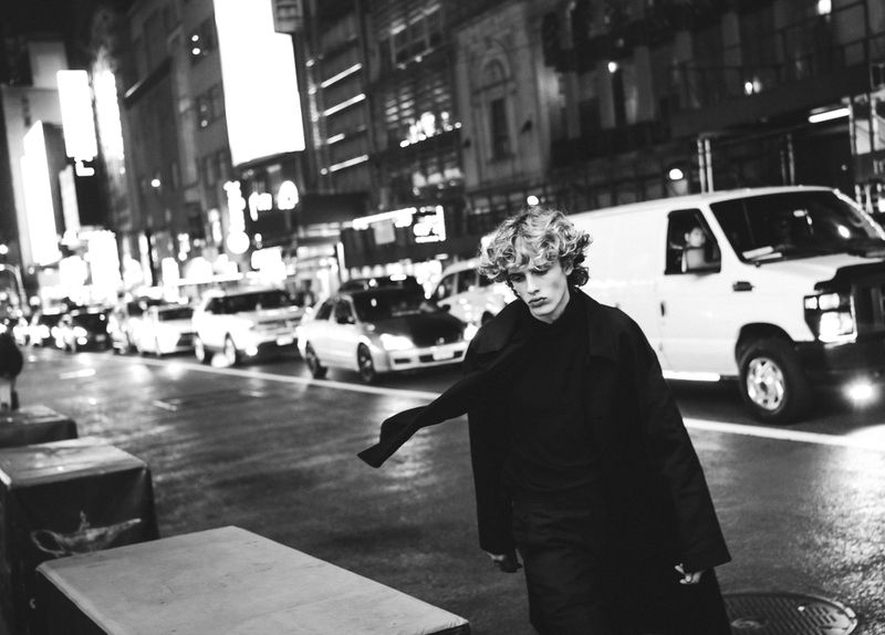 Monochrome night shot of a curly-haired man in a black coat walking on a city street lined with cars and bright building lights.