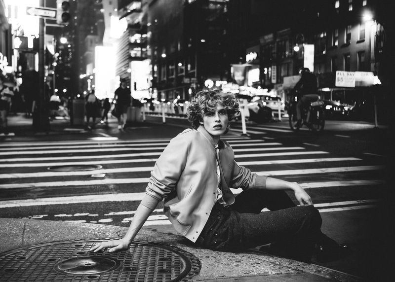 Model in a bomber jacket sits on a manhole cover in a crosswalk at night in an urban street scene. People and traffic in the background.