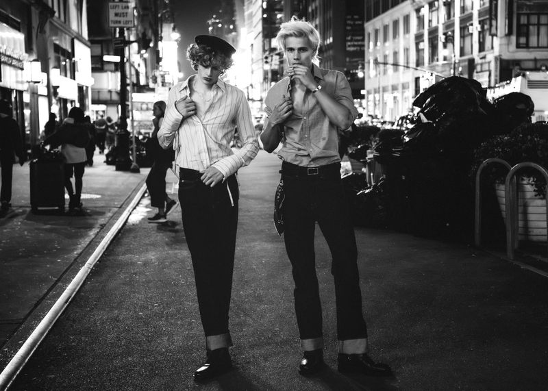 Black and white image of two people standing on a city street at night, wearing fashionable clothes and posing for a photo.